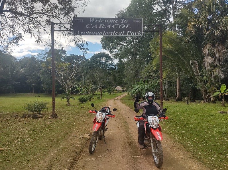 A woman on a motorcycle
        next to a riderless motorcycle, under a sign in a jungle
        regarding the ruins of Caracol in Belize