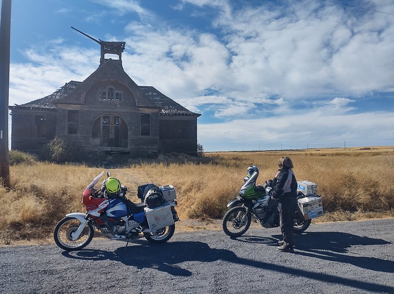 two motorcycles in front of
        an old schoolhouse that is falling apart. A woman stands looking
        at it with her back to the camera.